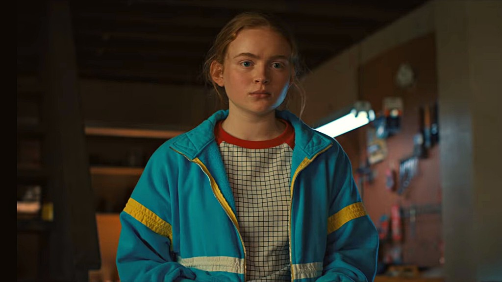 Max Mayfield con chaqueta azul de bandas amarillas y cuello rojo, en un taller